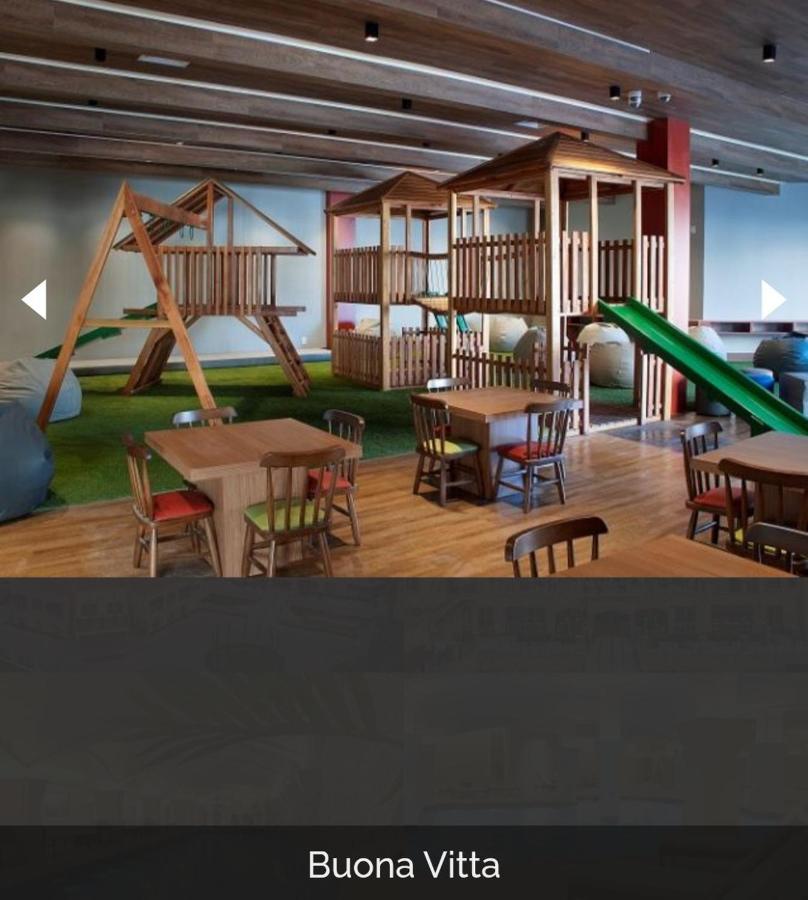 Buona Vitta Gramado Resort & Spa By Gramado Parks מראה חיצוני תמונה The photo shows a play area designed for children, featuring a wooden play structure with slides and climbing areas. In the foreground, there are several small wooden tables and chairs arranged for seating. The floor is covered with green turf, resem