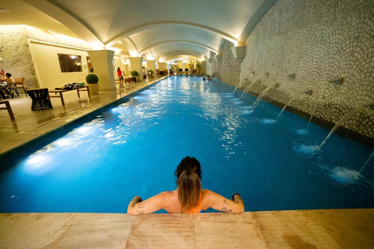 Buona Vitta Gramado Resort & Spa By Gramado Parks מראה חיצוני תמונה The photo shows an indoor swimming pool with a long, narrow design. The water is a clear blue, and there are features such as fountains along one side. In the foreground, a person with long hair is sitting on the edge of the pool, looking out over th