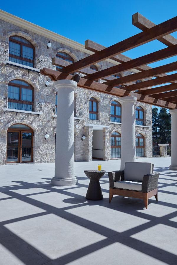 Buona Vitta Gramado Resort & Spa By Gramado Parks מראה חיצוני תמונה The photo shows a well-designed outdoor space with a stone wall building in the background. There are several tall columns that support a wooden pergola overhead, creating a shaded area. In this space, there is a stylish chair and a small table besid