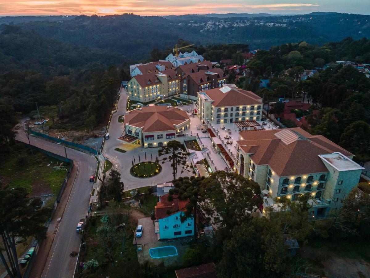 Buona Vitta Gramado Resort & Spa By Gramado Parks מראה חיצוני תמונה The photo shows an aerial view of a hotel complex set in a hilly, wooded area. The buildings feature a mix of architectural styles with several structures, including a central round pavilion. Surrounding the buildings are well-maintained gardens and 