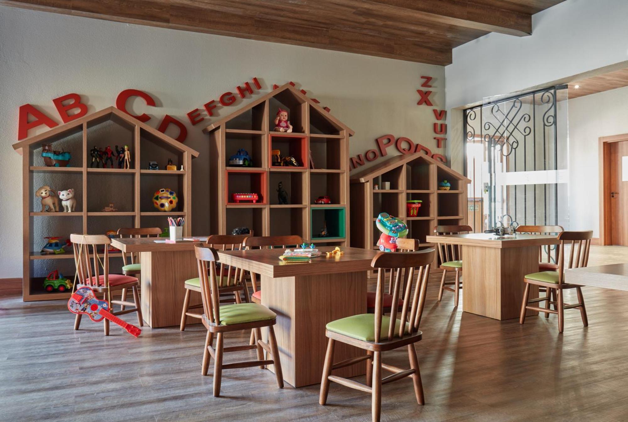 Buona Vitta Gramado Resort & Spa By Gramado Parks מראה חיצוני תמונה The photo shows a classroom or playroom designed for children. It features wooden furniture, including several tables and chairs with green cushions. In the background, there are large, open shelving units shaped like houses, displaying various toys 