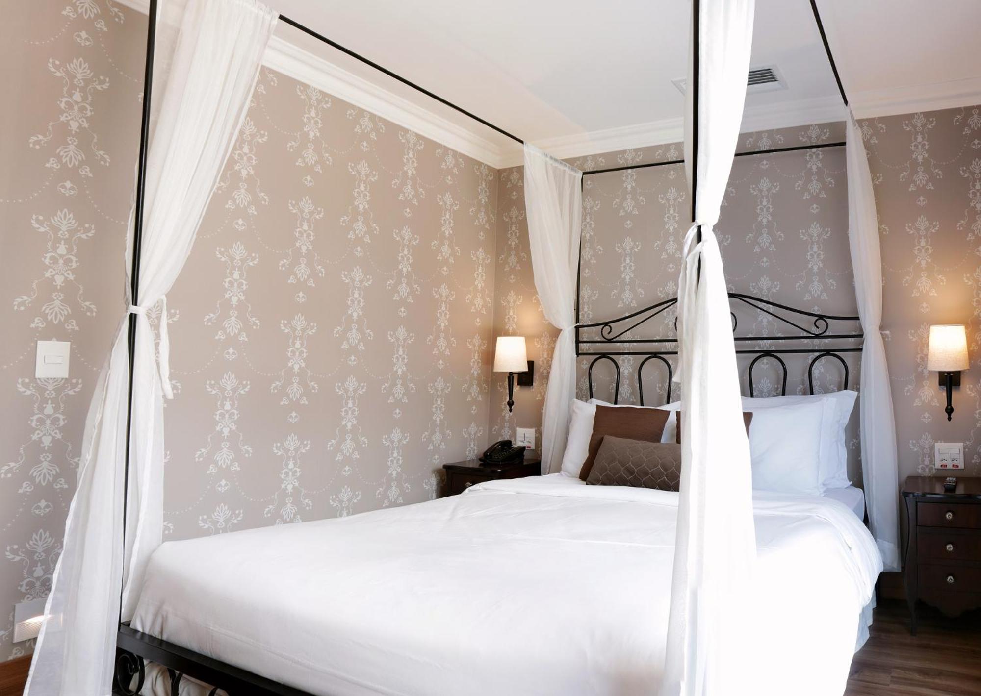 Buona Vitta Gramado Resort & Spa By Gramado Parks מראה חיצוני תמונה The photo shows a elegantly decorated bedroom featuring a four-poster bed with white drapes. The bed is made with white linens and has some brown accent pillows. The walls are adorned with a subtle floral pattern, and there are two bedside lamps prov