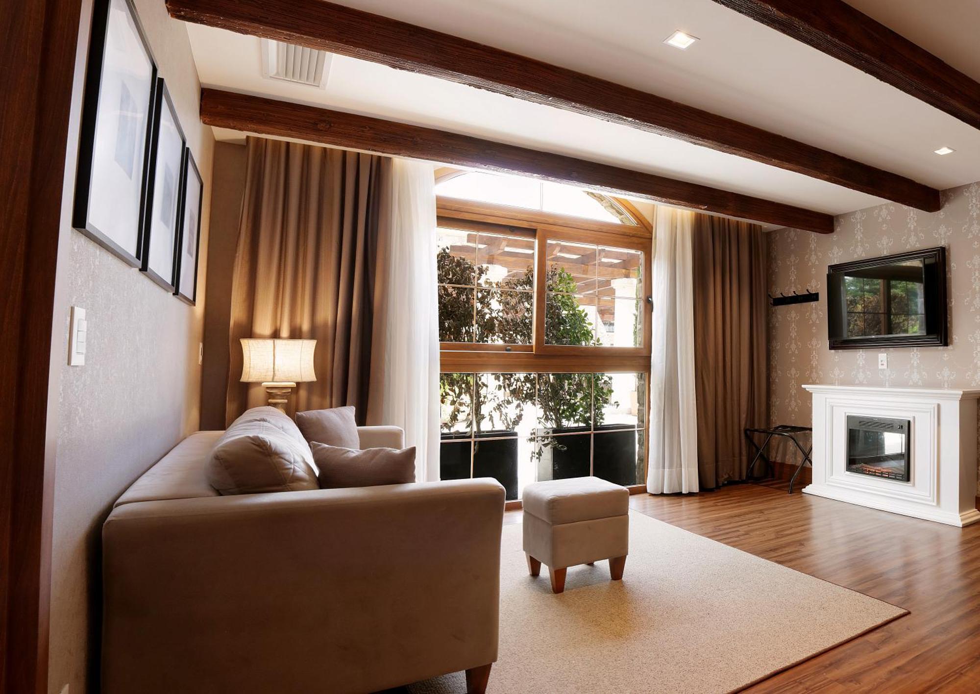 Buona Vitta Gramado Resort & Spa By Gramado Parks מראה חיצוני תמונה The photo depicts a cozy living room setting. It features a light-colored couch, a small ottoman, and a floor lamp beside the couch. Large windows are covered with curtains, allowing natural light to illuminate the space. In the background, there is 