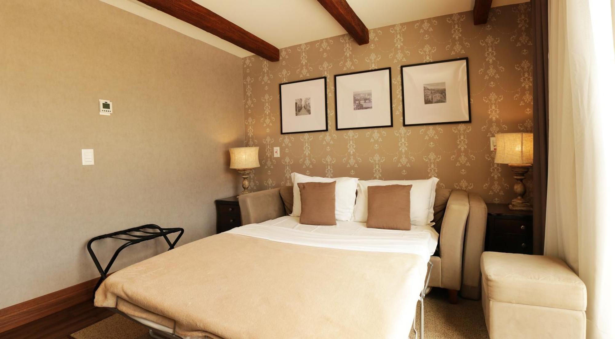 Buona Vitta Gramado Resort & Spa By Gramado Parks מראה חיצוני תמונה The photo shows a cozy bedroom with a neutral color palette. There is a bed with white linens and two brown decorative pillows. On either side of the bed, there are bedside tables with lamps. The walls feature a subtle floral pattern, and three frame
