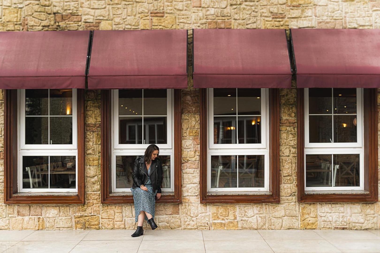 Buona Vitta Gramado Resort & Spa By Gramado Parks מראה חיצוני תמונה The photo shows a woman standing against a stone wall with three large windows in front of her, each framed with wooden borders. The windows have maroon awnings above them. The woman is dressed in a long skirt and leather jacket, leaning slightly aga